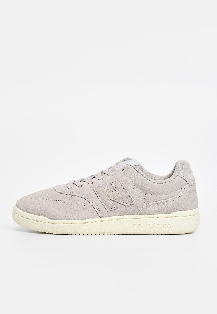 Zapatilla de ante gris claro con puntera redonda, cuello acolchado, cordones blancos, logo "N" prominente en el lateral y suela de goma crema.