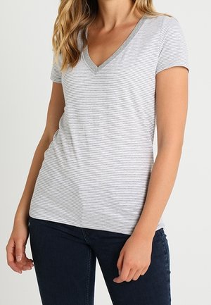 Vrouw die een aansluitend lichtgrijs-wit gestreept V-hals T-shirt draagt, gecombineerd met donkerblauwe spijkerbroek, staand met ontspannen armen.