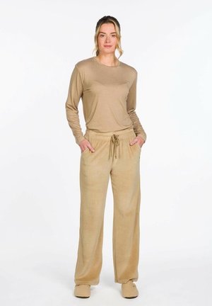 Frau steht mit den Händen in den Taschen, trägt ein beige langärmeliges Hemd, beige Hose mit Kordelzug und passende beige Slipper auf weißem Hintergrund.