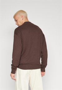 Jersey marrón de punto con cuello redondo, mangas largas y puños y dobladillo de canalé, combinado con pantalones sueltos de color claro.
