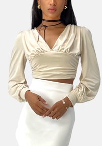 Beige satijnen cropped top met lange pofmouwen en een geraffineerd voorpand. Gecombineerd met een witte aangesloten rok. Zwarte choker en gouden oorbellen als accessoires.