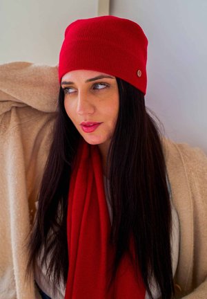 Rød strikket beanie med et tekstureret mønster og et lille metal-logo, parret med et rødt tørklæde og et lysfarvet sjal.