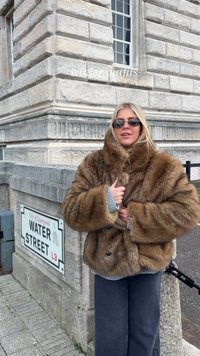 Bruine winterjas van kunstbont met een hoge kraag, oversized pasvorm en zwarte knopen. Gecombineerd met grijze geribbelde manchetten en donkere broek.