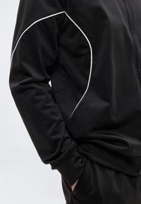 Μαύρο αθλητικό μπουφάν με φερμουάρ και λευκή διακοσμητική ραφή, λεία υφή, μακριά μανίκια, ελαστικές μανσέτες και ψηλό γιακά. Μινιμαλιστικός σχεδιασμός.