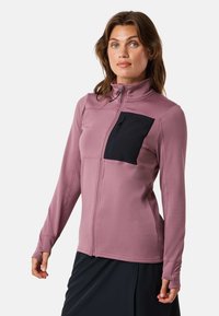 Donna che indossa una giacca mauve con zip integrale, tasca sul petto con zip nera, maniche lunghe e fori per i pollici, abbinata a una gonna nera.