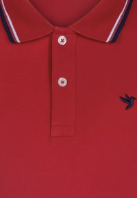 Polo rouge en tissu texturé, doté d'un col classique avec des accents rayés bleu marine et blanc et une fermeture à trois boutons. Logo d'oiseau bleu foncé.