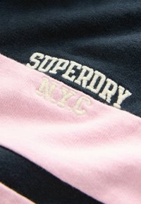 Marineblaue und rosa gestreifte Stoff mit gesticktem "SUPERDRY" und "NYC" in Weiß. Weiche Textur mit Kontrastnähten.