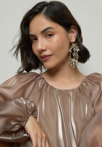 Gold floral chandelier earrings adorned with clear crystals, paired with a flowing, sheer taupe blouse featuring gathered sleeves.