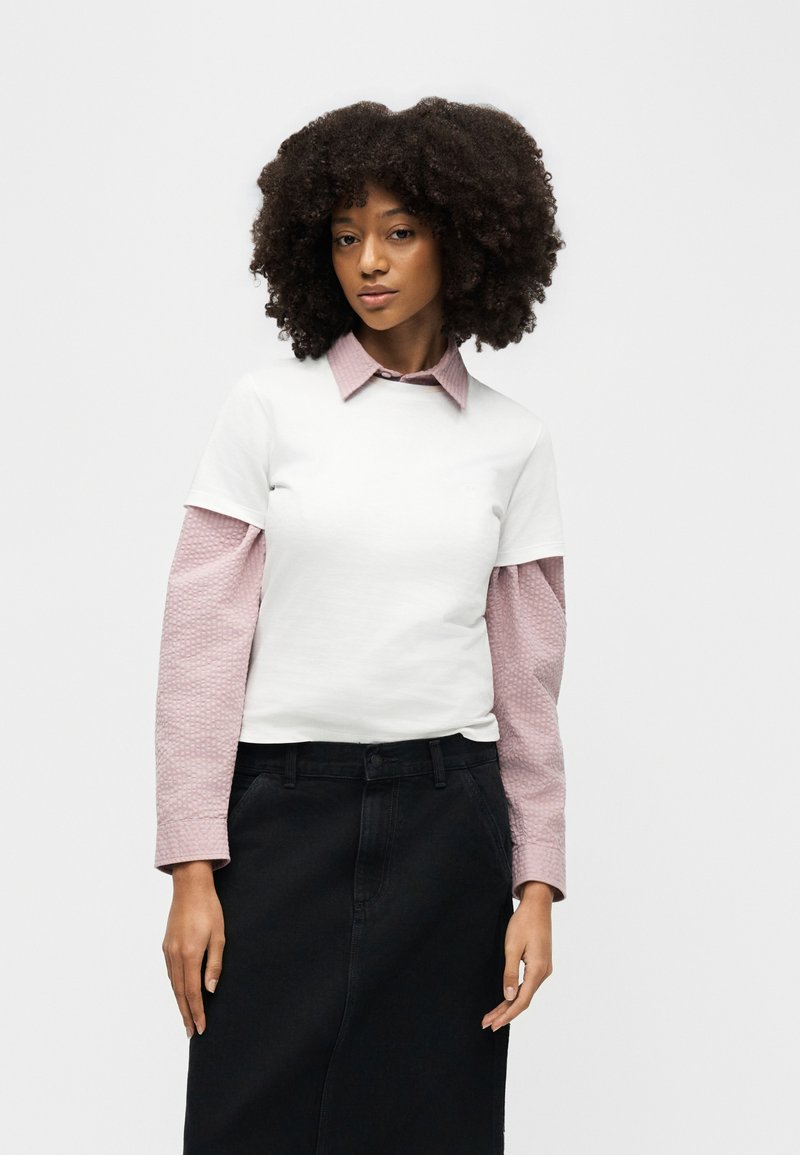 Giovane donna con capelli ricci che indossa una maglietta bianca a maniche corte sopra una camicia rosa a maniche lunghe con texture e una gonna nera.