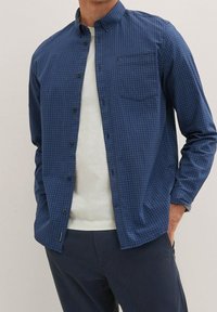 Camicia a quadri blu in un tessuto leggero con colletto aperto, taschino sul petto e chiusura con bottoni. Abbinata a un maglione leggero.