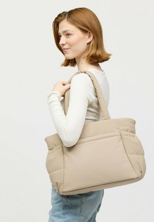 Jonge vrouw met rood haar draagt een grote beige gevoerde draagtas over haar schouder, gekleed in een wit shirt met lange mouwen en blauwe jeans, staand opzij.