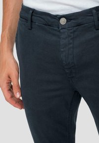 Chino in cotone blu scuro con una superficie liscia, dotato di chiusura anteriore con bottone e tasche laterali. Inquadratura ravvicinata della vita e della posizione della mano.