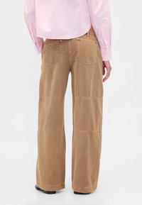 Pantalon cargo marron à jambes larges en coton résistant, avec des accents cousus et deux poches arrière doublées de surpiqûres décoratives.