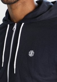 Felpa con zip color navy, dotata di lacci bianchi e realizzata in tessuto morbido. Presenta un ricamo a logo circolare sulla parte sinistra del petto.