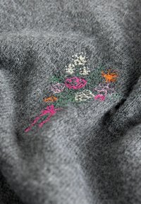 Pelēks audums ar teksturētu izskatu, kurā ir krāsaini ziedu izšuvumi rozā, zaļā, oranžā un baltā krāsā.