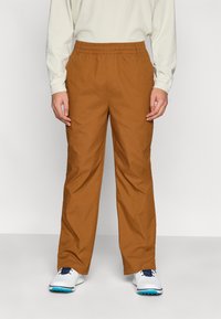 Pantalon marron à jambes droites avec une taille élastique. Fabriqué dans un tissu lisse et léger. Porté avec un haut crème à manches longues.