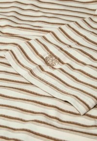 Tissu blanc avec des rayures horizontales marron régulièrement espacées et un petit blason brodé détaillé près du centre.