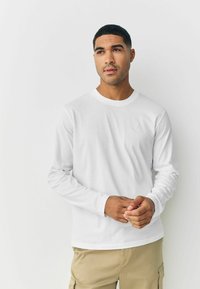 Long-sleeve white shirt made of cotton, featuring a round neckline and no visible patterns, paired with khaki pants.