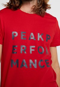 Camiseta de algodón roja con un atrevido texto gráfico en negro "PEAK PERFORMANCE". Diseño de cuello redondo con mangas cortas y textura suave.