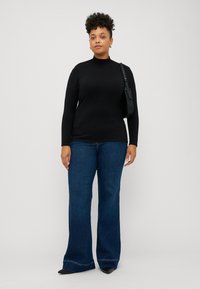 Haut noir à manches longues avec un col montant, associé à un jean en denim bleu à jambes larges. La tenue comprend un sac à main noir et des chaussures pointues noires.