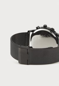 Bracelet de montre en mesh noir avec finition métallique, doté d'une fermeture à clips. Le cadran de la montre est rond et présente des éléments de chronométrage visibles.