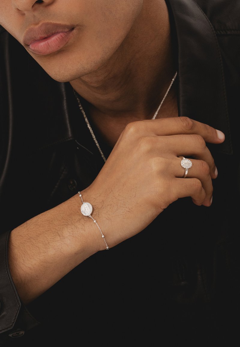 Gros plan d'une personne portant un bracelet et une bague en argent avec des pendentifs ovales, vêtue d'une veste en cuir noire et d'un collier.