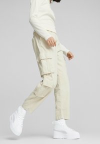 Pantaloni cargo beige con molte tasche laterali e una vestibilità comoda, abbinati a sneakers alte bianche con suola testurizzata e dettagli metallici.