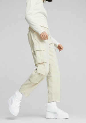 Pantalons cargo beiges avec plusieurs poches latérales et une coupe décontractée, associés à des baskets montantes blanches avec une semelle texturée et des accents métalliques.