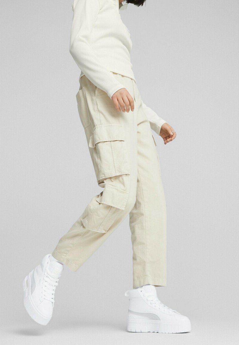 Pantaloni cargo beige con molte tasche laterali e una vestibilità comoda, abbinati a sneakers alte bianche con suola testurizzata e dettagli metallici.