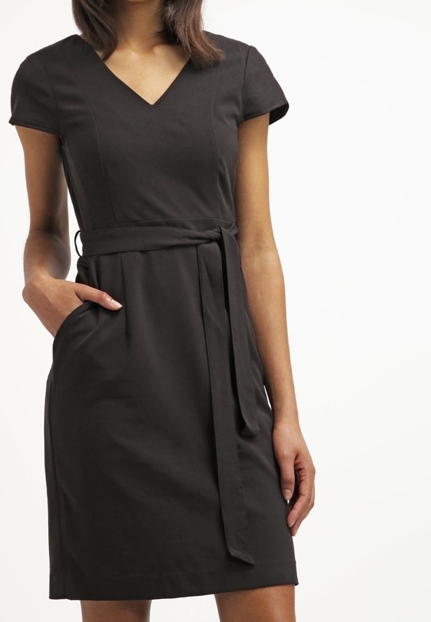 Femme portant une robe noire à manches courtes avec col en V et ceinture nouée à la taille, une main dans la poche, debout devant un fond clair uni.