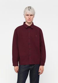 Bordeaux langærmet skjorte med knaplukning og spread-krave, med et diskret logo, sorte knapper og et glat, struktureret stof.