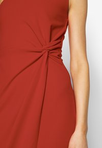 Röd klänning med knutdetalj i midjan, slät tyg och en figurnära silhuett. Texturen är mjuk och n ågot draperad.