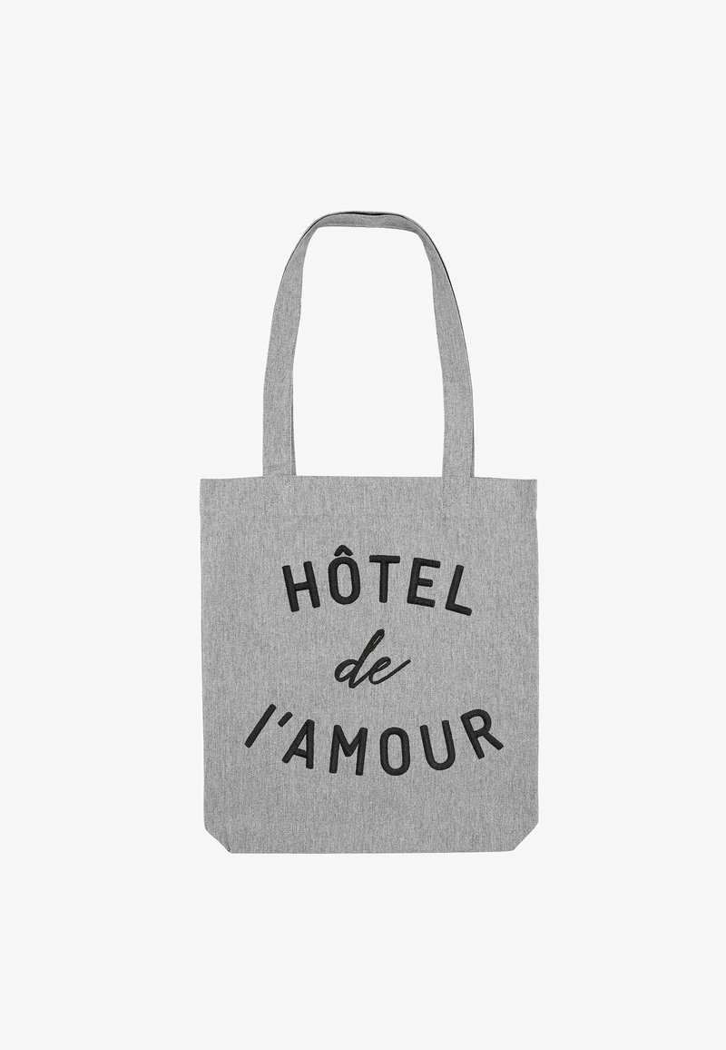 Sac fourre-tout en tissu gris avec deux poignées. Texte noir "HÔTEL de l'amour" imprimé en évidence sur le devant. Forme rectangulaire simple.