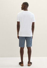 Weißes Baumwoll-T-Shirt, graue Shorts und braune Sandalen, von hinten vor einem hellen neutralen Hintergrund betrachtet. Einfaches Design, kurze Ärmel.