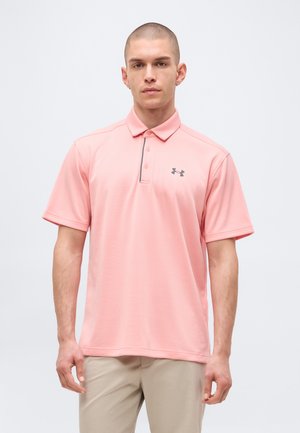 Muž s krátkymi vlasmi, ktorý nosí svetloružové polo tričko Under Armour a béžové nohavice, stojí pred jednoliatym svetlým pozadím.