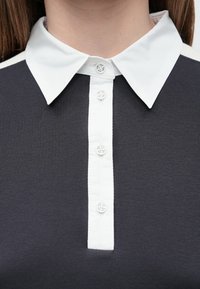 Detail bieleho košeľového goliera na tmavosivej rebrovanej košeli so štyrmi bielymi gombíkmi na prednom zapínaní.