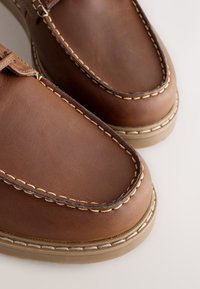 Mocassins en cuir marron avec coutures beige, bout arrondi et semelle en caoutchouc. Présentent une texture douce et des éléments de design classiques.