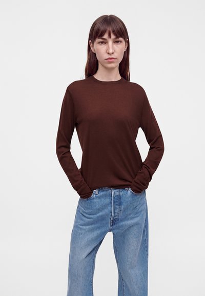 Γυναίκα με μακριά καστανά μαλλιά, που φοράει σκούρο καφέ μακρυμάνικο πουκάμισο και φαρδιά μπλε τζιν, στέκεται με τα χέρια στις τσέπες μπροστά σε απλό φόντο.