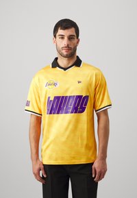 Gelbes Basketball-Trikot mit schwarzem Kragen, ausgestattet mit lila "LAKERS"-Schriftzug und Los Angeles-Logo. Kurze Ärmel und strukturierter Stoff.