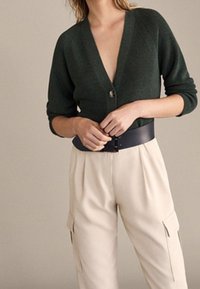 Gilet tricoté vert foncé avec un décolleté en V et une fermeture à un seul bouton, associé à un pantalon crème avec poches cargo et une large ceinture.