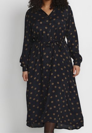 Woman wearing a long-sleeve black dress with brown polka dots, buttoned front, and tied waist.