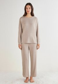 Lys beige loungewear sæt bestående af en langærmet top og løstsiddende bukser lavet af blødt stof, med subtile stikninger ved kanterne.