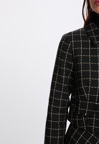 Cappotto nero a quadri con motivo a griglia bianca, caratterizzato da un tessuto strutturato, scollo con colletto e design corto.