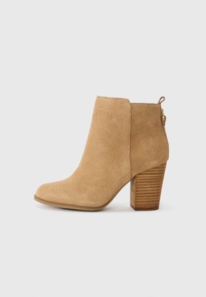 Bottines en daim beige avec un bout arrondi, un talon en bois texturé et une fermeture éclair sur le côté pour un enfilage facile. Fini lisse avec des coutures minimales.