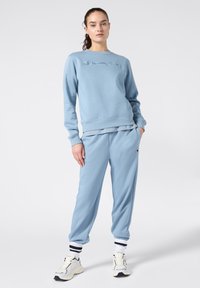 Una donna in piedi indossa un felpa Champion azzurro chiaro abbinato a pantaloni jogger, calzini bianchi con strisce nere e sneaker bianche su uno sfondo semplice.
