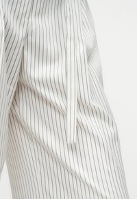 Λευκό ύφασμα με λεπτές μαύρες κάθετες ρίγες και κρεμαστό υφασμάτινο ζώνη ή κορδέλα στο πλάι, που δείχνει τη λεπτομέρεια της μέσης του ενδύματος.