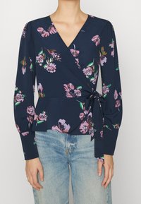 Top avvolgente floreale navy con maniche lunghe, caratterizzato da motivi floreali rosa e verdi, scollo a V profondo e una cintura da annodare in vita. Abbinato a jeans di denim.