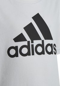 Camiseta de algodón blanca con un gran logo negro de Adidas, acompañado de tres franjas diagonales. Diseño de cuello redondo estándar y mangas cortas.