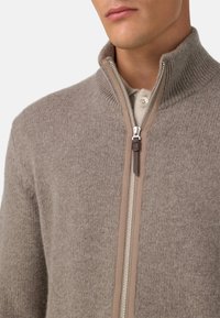 Beige svetr z vlněné směsi s vysokým límcem, ribkovanou strukturou a předním zipem s hnědým akcentem. Obsahuje zapínací vnitřní vrstvu.