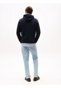 Felpa con cappuccio blu navy con vestibilità comoda e polsini a coste, indossata sopra una camicia a righe azzurre e bianche. Abbinata a jeans azzurri e scarpe scure.